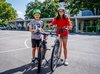 Une écolière et un écolier se tiennent dans la cour de récréation avec leur vélo et regardent l’objectif en souriant…..